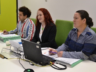 3 people at a desk with documents and a laptop. They are listening to someone.